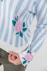 A close-up of a person wearing a white and light blue striped knit sweater with pink rose and green leaf motifs on the front and cuff, paired with olive green pants.