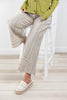 A person seated on a white stool wearing wide-leg striped pants in beige, cream, and muted blue tones, with a green cardigan and cream loafers in a bright indoor room.