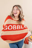 A woman wearing a chunky knit sweater with bold horizontal stripes in terracotta orange and cream, featuring the word 'GOBBLE' in large white letters across the chest. She is also wearing blue jeans and gold hoop earrings, standing against a solid beige background.