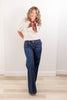 A woman modeling a cream puff-sleeve blouse with a red and leopard-print neck scarf, paired with dark blue flare jeans, standing in a bright room with light wood floors and white walls.