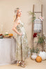 A woman models a sleeveless, midi-length dress with a beige and green botanical pattern, standing in a styled fall-themed room with pumpkins, a ladder, and a table.
