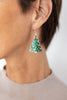 A close-up of a woman's ear wearing a dangling earring shaped like a Christmas tree, with green enamel, small colorful ornaments, and a gold star on a silver hook.