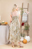 A woman stands with her back to the camera wearing a sleeveless midi dress with a patchwork floral print in muted beige, sage green, and brown tones. The dress features a smocked bodice and thin shoulder straps.