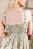 A woman shown from the back wearing a sleeveless smocked dress with a muted floral and paisley print in beige, sage green, and brown tones. The dress features thin straps with a patterned trim and small metal O-ring details.