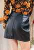 A close-up of a person wearing a black faux-leather mini skirt with a back zipper, paired with a black floral long-sleeve top.