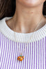 A close-up of a person wearing a gold chain necklace with a small orange jack-o'-lantern pendant and a tiny matching pumpkin charm, styled over a purple and white striped knit sweater.