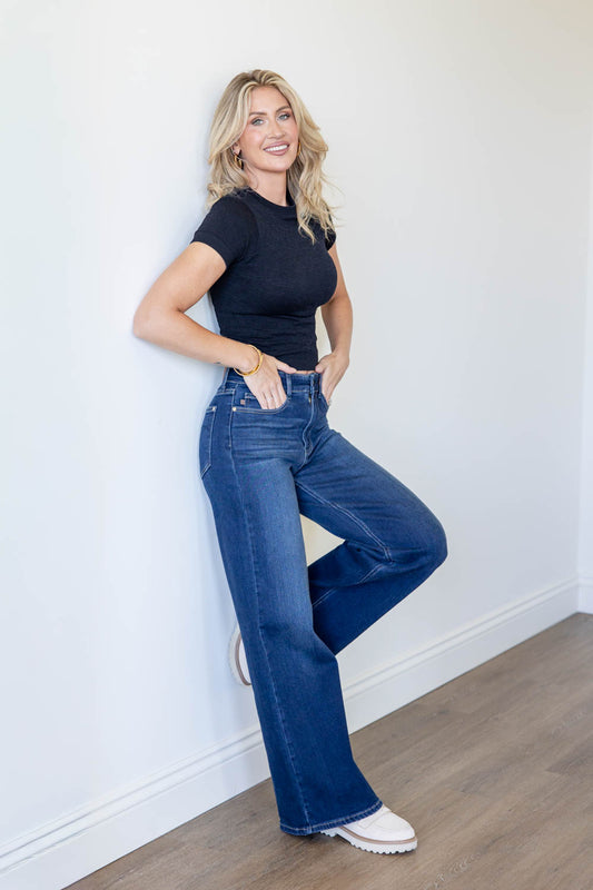 A woman posing in a black short-sleeve t-shirt and high-waisted, wide-leg blue jeans against a white wall with light wood flooring.
