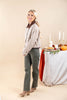 A woman models a beige and white gingham cropped jacket with a brown collar, paired with olive green jeans and beige ankle boots, standing in front of a neutral beige background with a styled autumn table in the background.