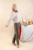 A woman modeling a cropped beige and white gingham button-up jacket with a brown collar, paired with olive green pants and tan boots, standing next to a styled autumn table.