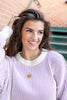 A smiling young woman with wavy brown hair wears a lavender and white vertically striped knit sweater with a cream ribbed collar. She is wearing a small orange pumpkin pendant necklace and posing in front of a brick wall and green steps.