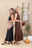 Two women modeling long sleeveless dresses in a styled studio setting. One dress is black with a brown and white striped scarf tied around the waist, and the other is a solid brown textured dress.