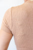 Close-up of a person wearing a short-sleeve knit top with a floral lace pattern in a light peach-beige color, shown from the back against a plain white background.