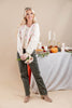 A woman stands in a studio setting wearing a cream knit sweater with pink floral embroidery, dark olive green pants, and taupe ankle boots. She is holding a small bouquet of flowers, with a styled autumn table backdrop featuring candles, a pumpkin, and greenery behind her.