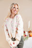 A smiling blonde woman wearing a cream knit sweater with pink floral embroidery down the front, paired with gray pants, standing in a warmly lit indoor setting with candles and autumn decor in the background.