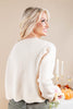 A woman with blonde hair is shown from the back wearing a cream-colored knit sweater and olive green pants, standing in front of a warm beige background with decorative pumpkins and candles.
