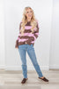 A woman modeling a pink and brown striped knit sweater, paired with light blue jeans and brown slip-on shoes, standing against a white wall and light wood floor.