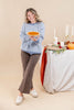 A woman stands in a studio setting holding a pumpkin pie, wearing a light blue and white scalloped sweater, brown flared pants, and white loafers. To her right is a decorated table with white candles, autumn greenery, and a small pumpkin against a warm beige backdrop.