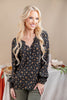 A smiling blonde woman wearing a black long-sleeve blouse with a small orange and yellow floral pattern, paired with olive green pants, standing in front of a beige background with a decorated table.