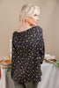 A woman with blonde hair is shown from the back wearing a black long-sleeve blouse with a small orange and yellow floral pattern. She is standing in front of a softly lit beige background with a decorated table visible behind her.