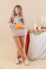 A woman stands in a studio setting wearing a beige knit cardigan with brown and orange flower patches, tan pants, and white shoes, while holding an orange pumpkin. A decorated table with candles, a pie, and autumn greenery is visible to her right against a warm beige backdrop.