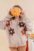 A person wearing a beige knit cardigan with brown and orange flower appliqués, holding a small orange pumpkin in front of their face against a warm beige background.