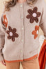 A close-up of a person wearing a beige knit cardigan with large brown and orange flower appliqués and cream buttons, paired with tan pants.