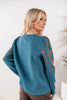 A woman wearing a teal blue knit sweater with pink floral and gold vine embroidery on the sleeves, shown from a back-three-quarter view against a bright white indoor background.
