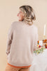 A woman with blonde hair is shown from behind wearing a light beige knit sweater with a contrasting rust-orange ribbed hem, standing in front of a warm beige background.