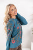 A woman wearing a teal long-sleeve sweater with pink floral and gold vine embroidery, paired with brown pants.