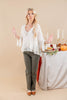 A woman modeling a white tiered blouse and olive green pants stands next to a decorated table with candles, autumn leaves, and pumpkins against a solid beige background.