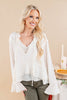 A blonde woman wearing a white long-sleeve blouse with a V-neck, lace trim, and flared cuffs, paired with olive green pants, posed against a warm beige background.