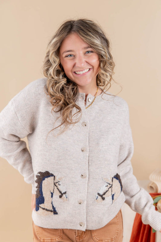 A smiling woman with wavy blonde hair wears a light beige knit cardigan featuring embroidered horse designs on the lower front, paired with tan pants, against a solid beige background.