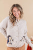 A smiling woman with wavy blonde hair wears a light beige knit cardigan featuring embroidered horse designs on the lower front, paired with tan pants, against a solid beige background.