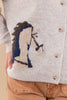 Close-up of a woman wearing a light beige knit cardigan with a knitted horse design in navy blue, brown, and cream, paired with tan pants.
