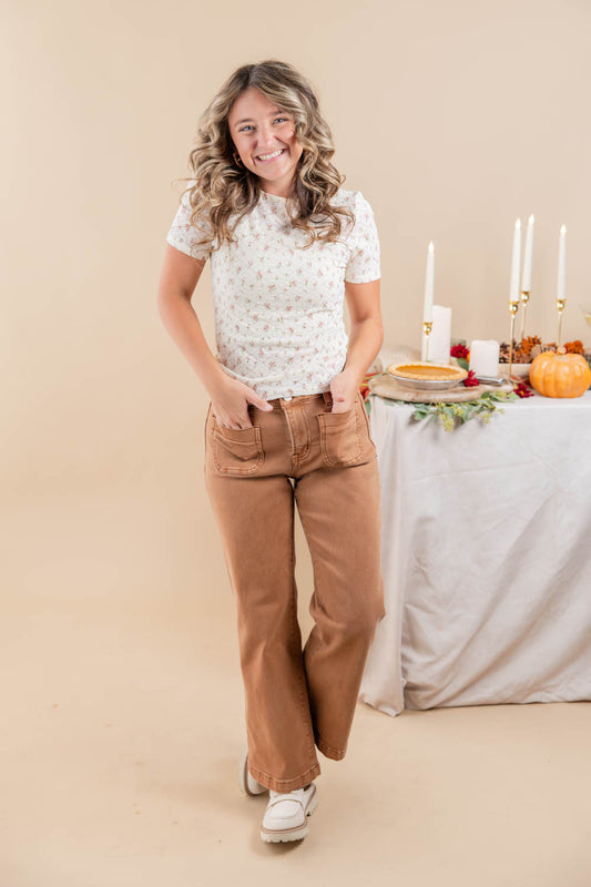 A woman modeling a white short-sleeve t-shirt with a small yellow floral pattern and brown wide-leg jeans, standing in front of a beige studio backdrop with a decorated autumn table in the background.