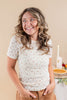 A smiling woman with wavy brown hair wearing a short-sleeve cream t-shirt with a tiny pink and green floral pattern, paired with brown pants, standing in a warmly lit indoor setting.