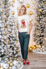A woman stands wearing a cream short-sleeve top with a sitting dog graphic, dark teal pants, and red heels.