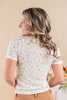 A woman with long wavy blonde-highlighted hair is shown from the back, wearing a short-sleeve cream-colored t-shirt with a small pink and green floral pattern, paired with tan pants.