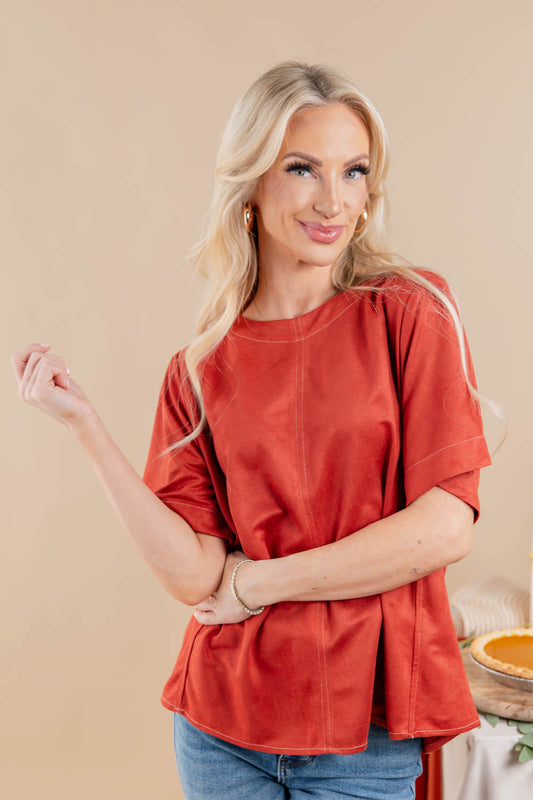 A blonde woman wearing a rust-orange faux suede short-sleeve top and blue jeans, posing against a solid beige background.