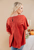 A woman shown from the back wearing a rust-orange faux suede short-sleeve top with a round neckline and a small back slit, paired with blue jeans in an indoor styled setting.