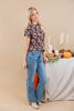 A woman modeling a brown short-sleeve top with a white floral pattern and light blue high-waisted flare jeans, standing next to a decorated table with candles, a pumpkin, and a pie against a beige studio background.