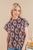 A woman wearing a brown short-sleeve top with a white floral pattern, paired with blue jeans, standing against a solid beige background.
