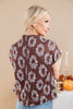 A woman shown from the back wearing a brown short-sleeve top with a white floral wreath pattern, paired with light blue jeans, set against a warm beige background with a decorative autumn table in the distance.
