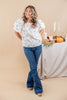 A woman modeling a white short-sleeve blouse with a delicate green floral pattern and puffed sleeves, paired with blue flare jeans, standing in front of a beige studio backdrop with a styled autumn table in the background.