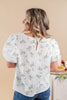 A woman shown from the back wearing a white textured blouse with a green and white floral print, puff sleeves, and a small keyhole cutout at the neckline, paired with blue jeans against a warm beige background.