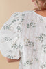 Close-up of a woman wearing a white textured blouse with green floral embroidery and vertical embroidered stripes, against a warm beige background.