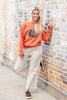 A woman posing against a brick wall wearing an orange crewneck sweatshirt with a plaid pumpkin graphic and cream pinstripe cropped pants.