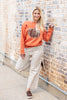 A woman posing against a brick wall wearing an orange crewneck sweatshirt with a plaid pumpkin graphic and cream pinstripe cropped pants.