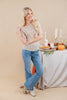 A woman modeling a beige short-sleeve top with small black polka dots, paired with blue jeans and beige ankle boots, standing next to a decorated table with candles, a pumpkin, and a pie.