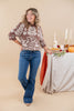 A woman modeling a brown and white patterned knit top with blue jeans, standing in a studio setting next to a decorated autumn table with pumpkins, candles, and a pie dish.